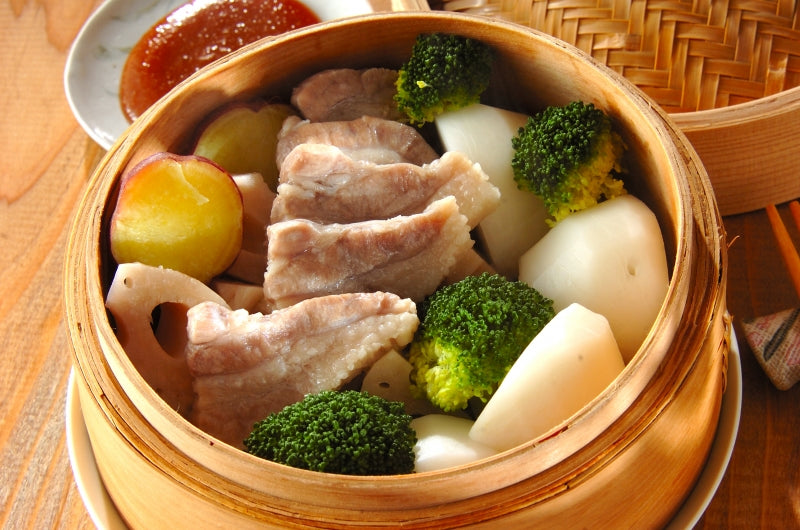 A bamboo steamer containing a variety of steamed foods including pork slices, broccoli, radish, and potato, with a side of sauce.