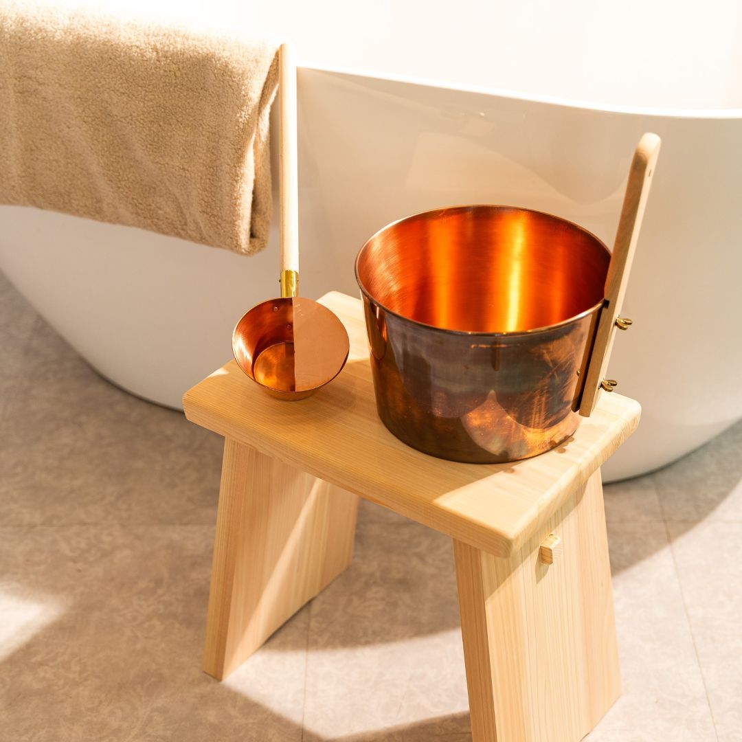 Japanese handmade copper sauna bucket with beechwood handle and matching copper ladle, placed on a natural hinoki wood stool beside a bathtub.