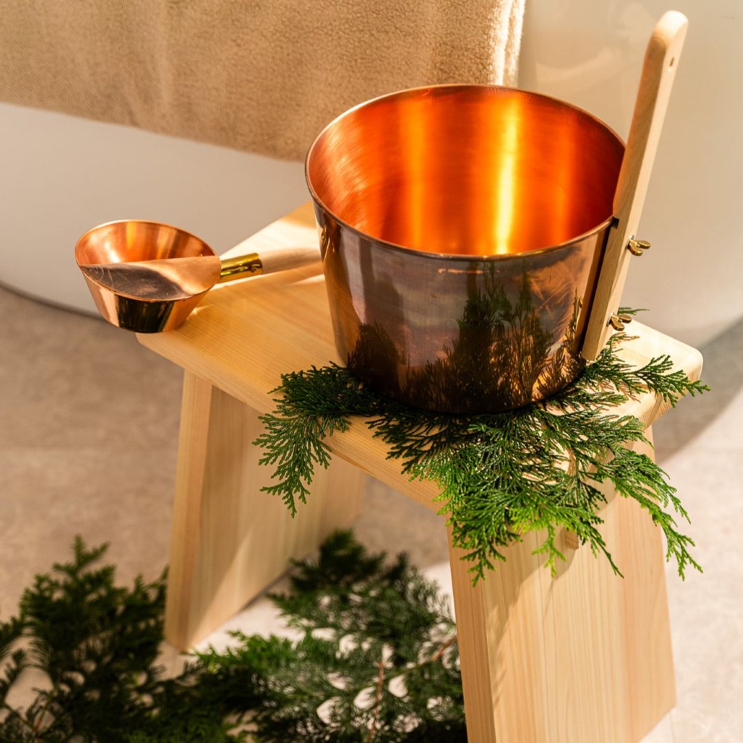 Copper sauna bucket and ladle on a hinoki wood stool decorated with fresh green cedar leaves, creating a natural spa atmosphere.