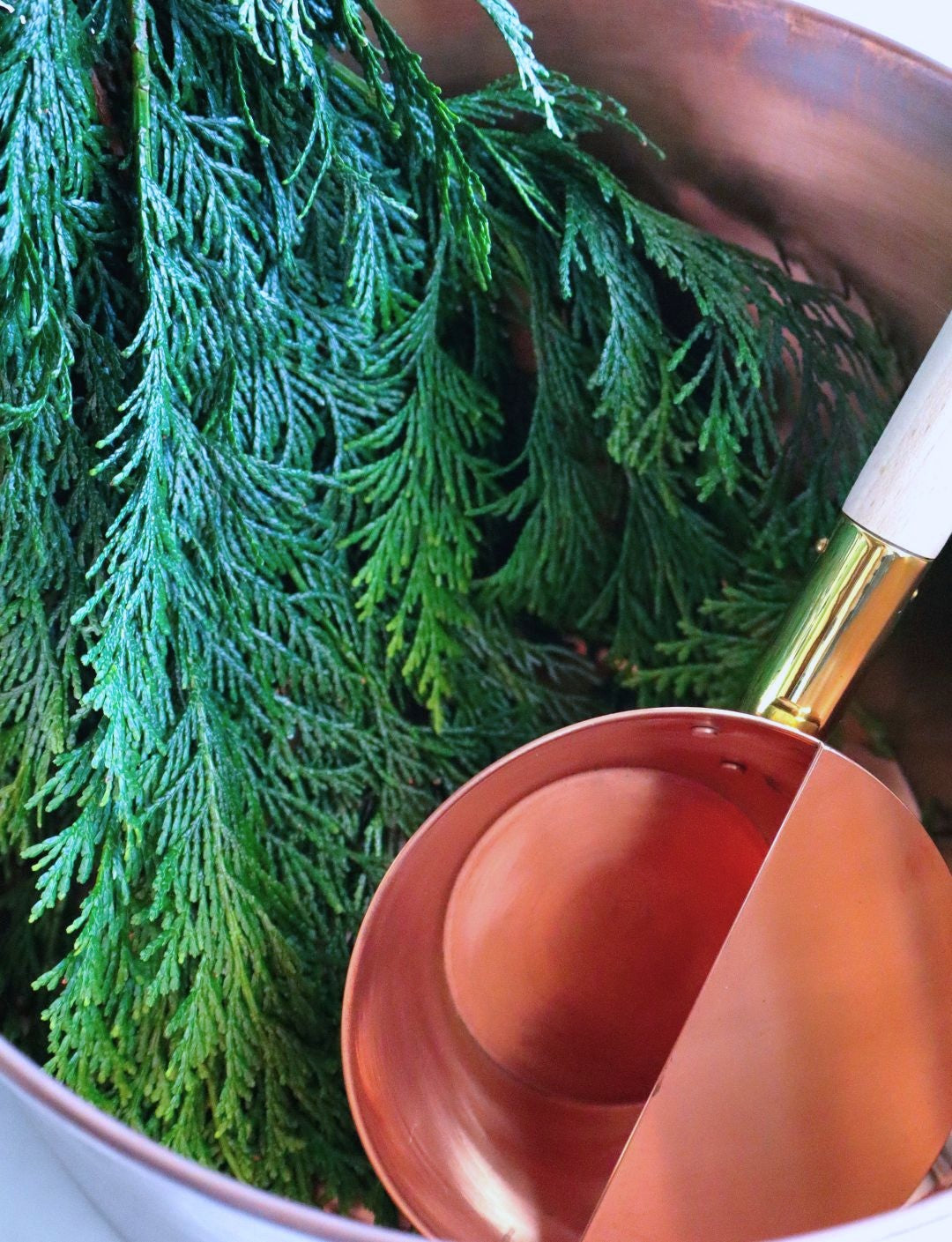 A Japanese copper sauna ladle placed inside a copper bucket with fresh hinoki branches, evoking a natural and refreshing sauna ambiance.