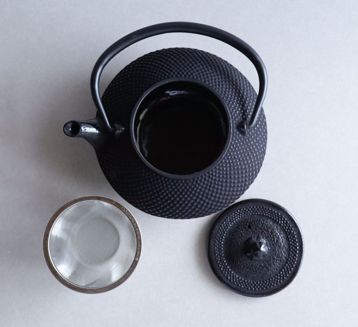 Top view of an open black Arare Nambu cast iron teapot with its lid removed and a stainless steel tea strainer placed beside it on a light surface.