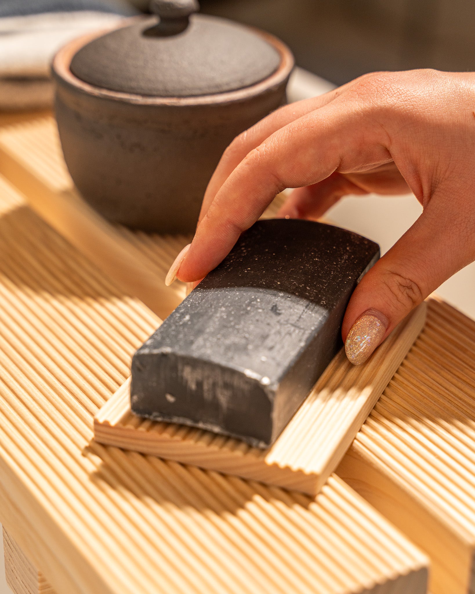 Hand holding a block of charcoal on a wooden surface with a teapot in the background
