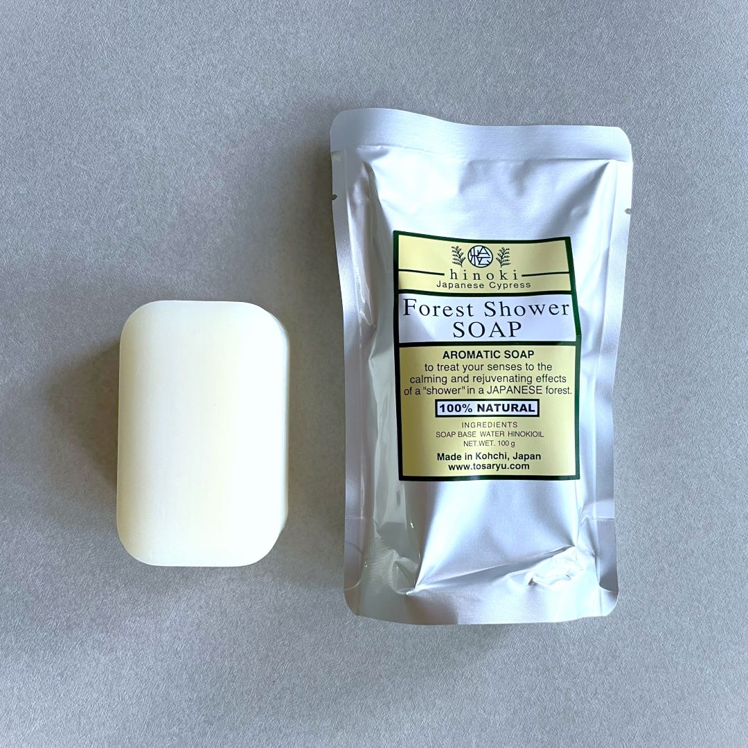 A bar of Hinoki Forest Shower Soap accompanied by its packaging. The soap bar is white and rectangular with rounded edges, while the packaging is a silver foil pouch labeled &