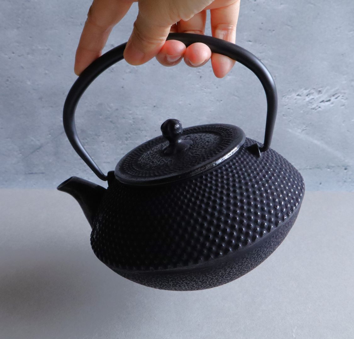 Hand lifting a black Arare Nambu cast iron teapot by its curved handle, showing the teapot&