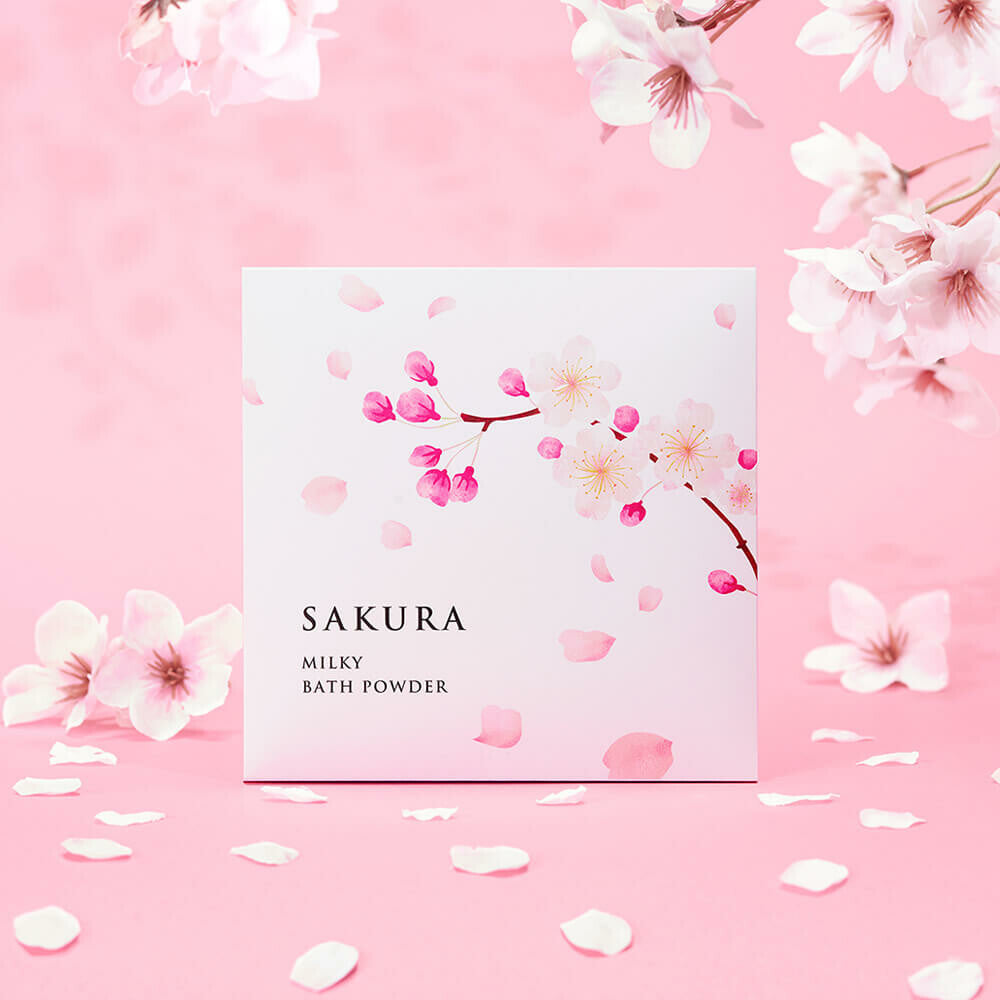 A beautifully packaged Sakura Milky Bath Powder with a delicate cherry blossom illustration on the front. The package is set against a pink background with scattered sakura petals, creating a soft and elegant Japanese aesthetic. Perfect for a luxurious bath experience infused with the gentle fragrance of cherry blossoms.