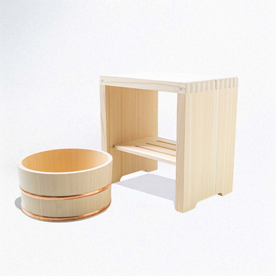 A wooden bucket next to a wooden stool in a bath room with a bath on background with green plants at the corner