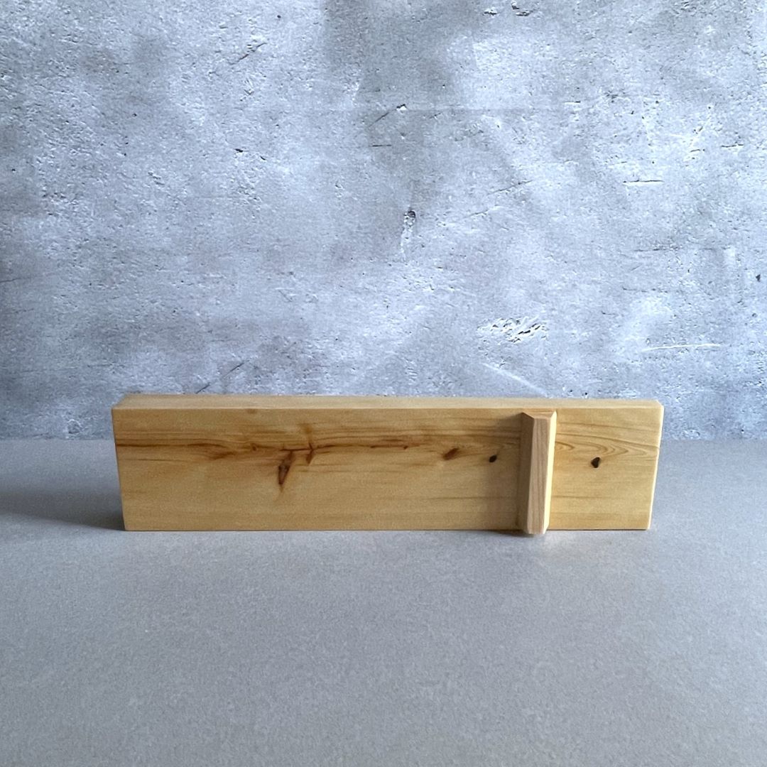 A wooden shelf with a natural finish and distinctive wood knots is placed horizontally in front of a textured concrete wall. The shelf is supported by a small wooden bracket at one end, creating a clean and functional design element within a minimalist setting.