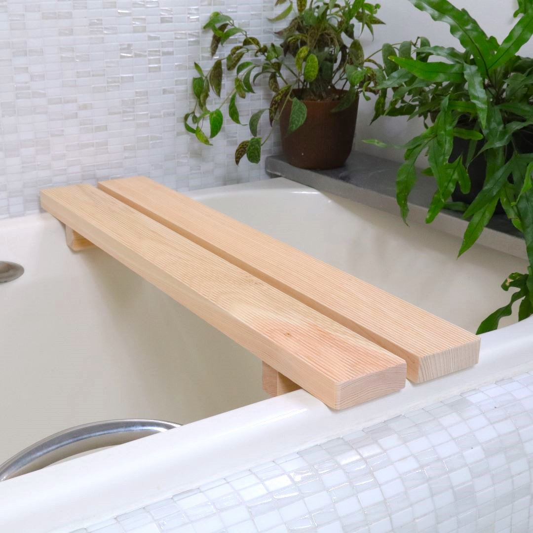 A light-hued Hinoki Cypress bathtub bench, also serving as a tub caddy, rests across a bathtub. At 27 inches long, it's perfectly sized for tubs between 20 to 23.6 inches wide, shown in a serene bathroom with a potted plant on a ledge.