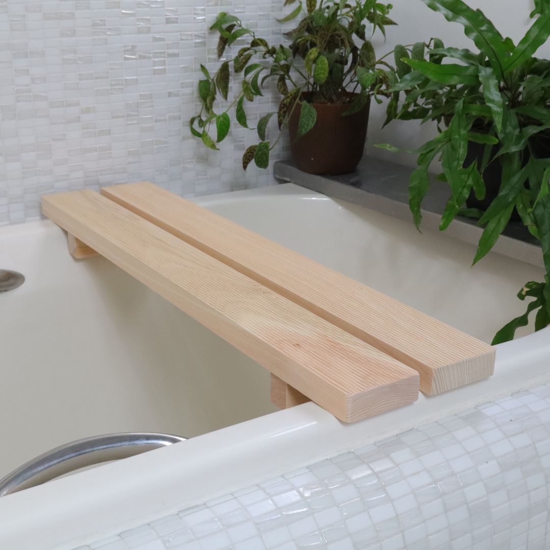 A minimalist Hinoki wood bath bench stretches across a white bathtub, juxtaposed against a mosaic tile backdrop with a lush potted plant, embodying a clean and tranquil spa setting.