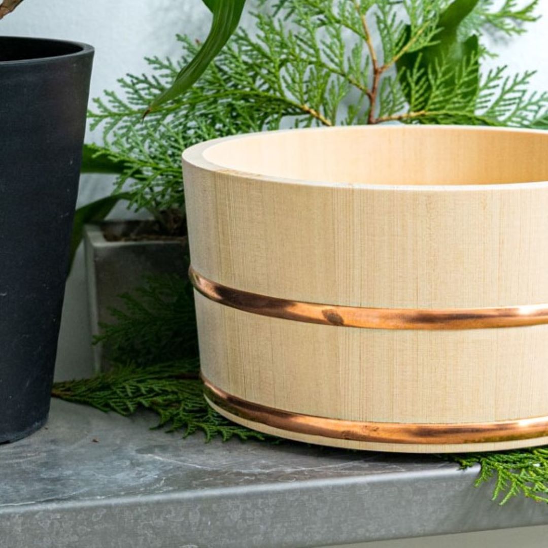 A bath wooden bucket with two copper ring around it a white towel on the edge and it is placed on a granit rock with the sea on background