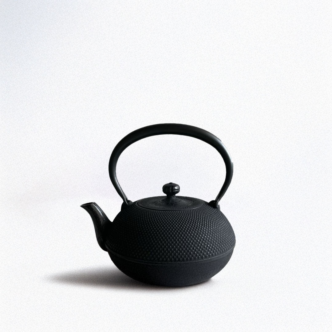 This is a black cast iron Tetsubin kettle with a lightly rough surface on the body. The iron handle has a smooth surface and is standing upright. The kettle is placed in the center of a space with grey walls and a grey surface, seen from a front view.