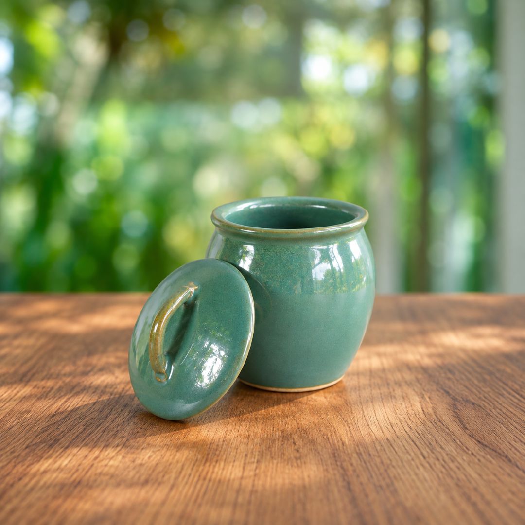 A small glazed ceramic jar with its lid leaning beside it on a wooden table, set against a softly blurred green outdoor background.