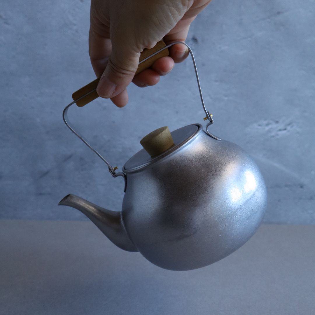Hand holding a silver teapot with a wooden handle against a neutral background