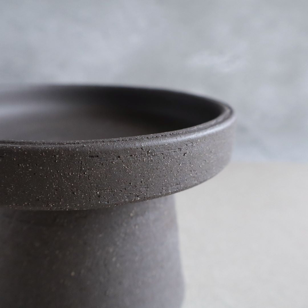 Close-up of the edge of a minimalist black ceramic footed bowl, highlighting the textured surface and the simple yet elegant design of the pottery against a soft-focus grey background.