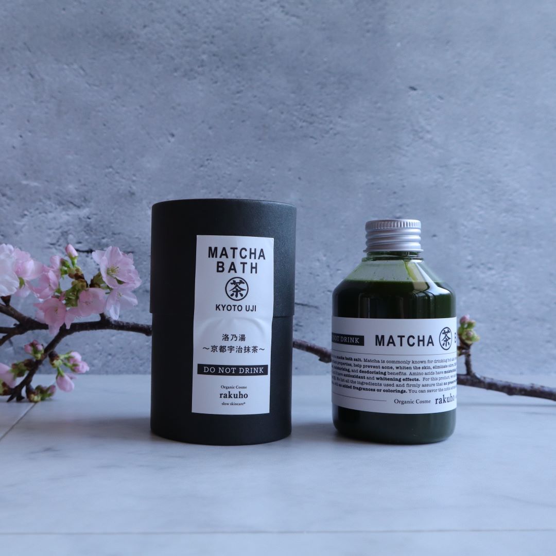 A bottle of Kyoto Uji Matcha Bath Lotion by "rakuho" stands next to its elegant cylindrical packaging, both placed on a marble surface with blooming sakura (cherry blossom) branches. The background is a soft gray wall, creating a minimalist, serene atmosphere that highlights the product’s refined and natural Japanese aesthetic.