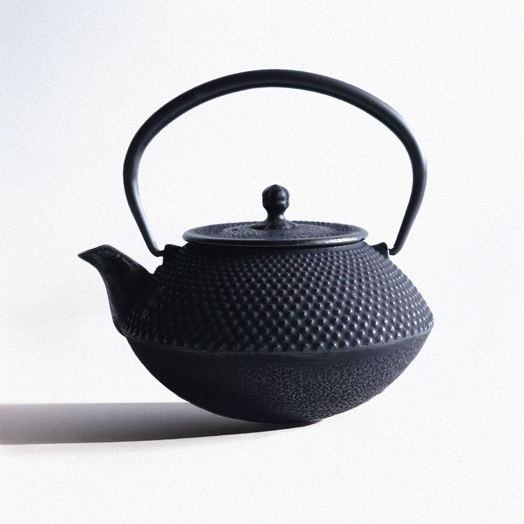 Black cast iron Japanese teapot in Arare Nambu style, featuring a textured dotted surface and curved handle, placed on a light grey surface with a soft concrete background.