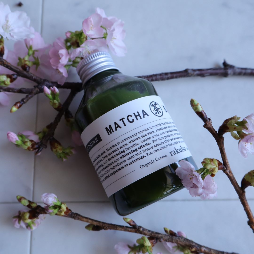 A glass bottle of matcha bath lotion from the brand "rakuka" rests elegantly among blooming sakura (cherry blossom) branches on a white tile surface. The green liquid contrasts beautifully with the delicate pink petals, evoking a serene, natural spa ambiance.