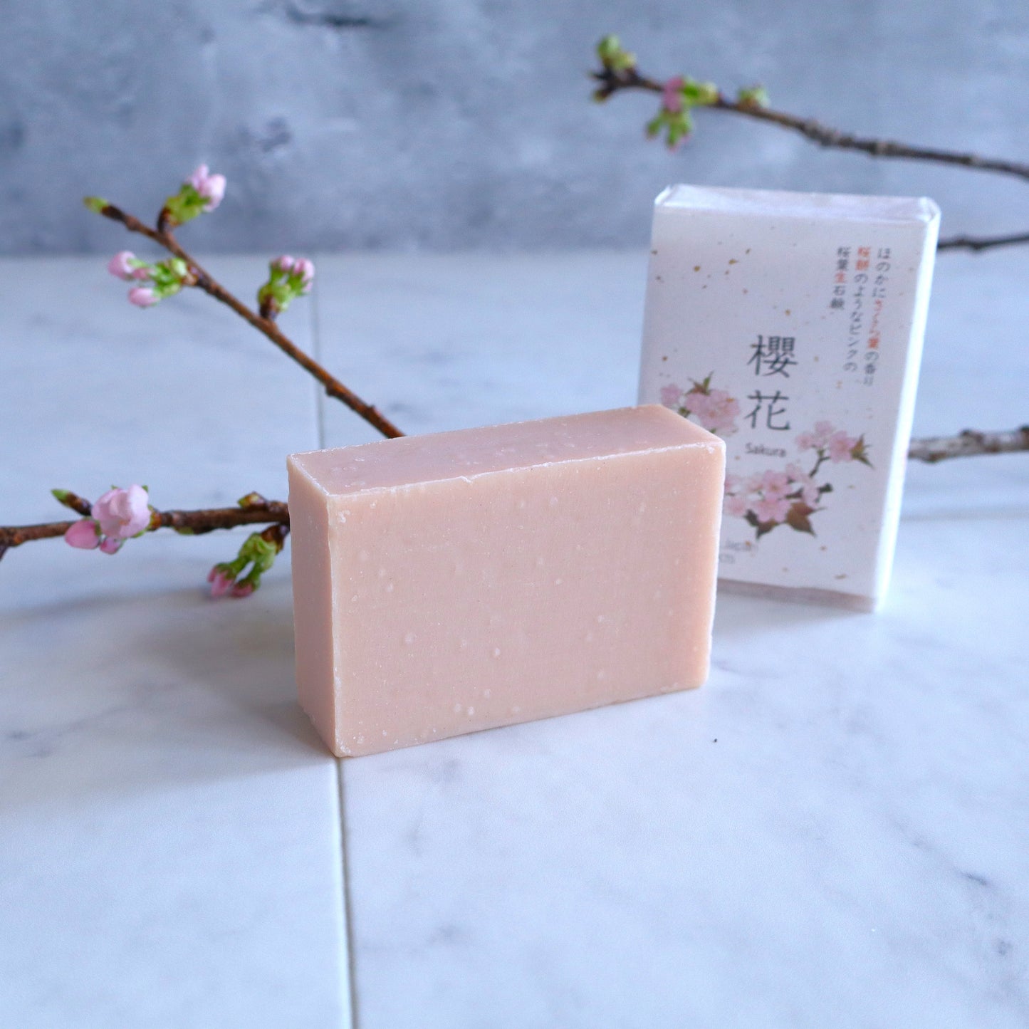 Japanese Sakura soap with a delicate pink hue, displayed alongside its elegant cherry blossom packaging on a marble surface, with blooming sakura branches in the background for a natural and serene aesthetic.