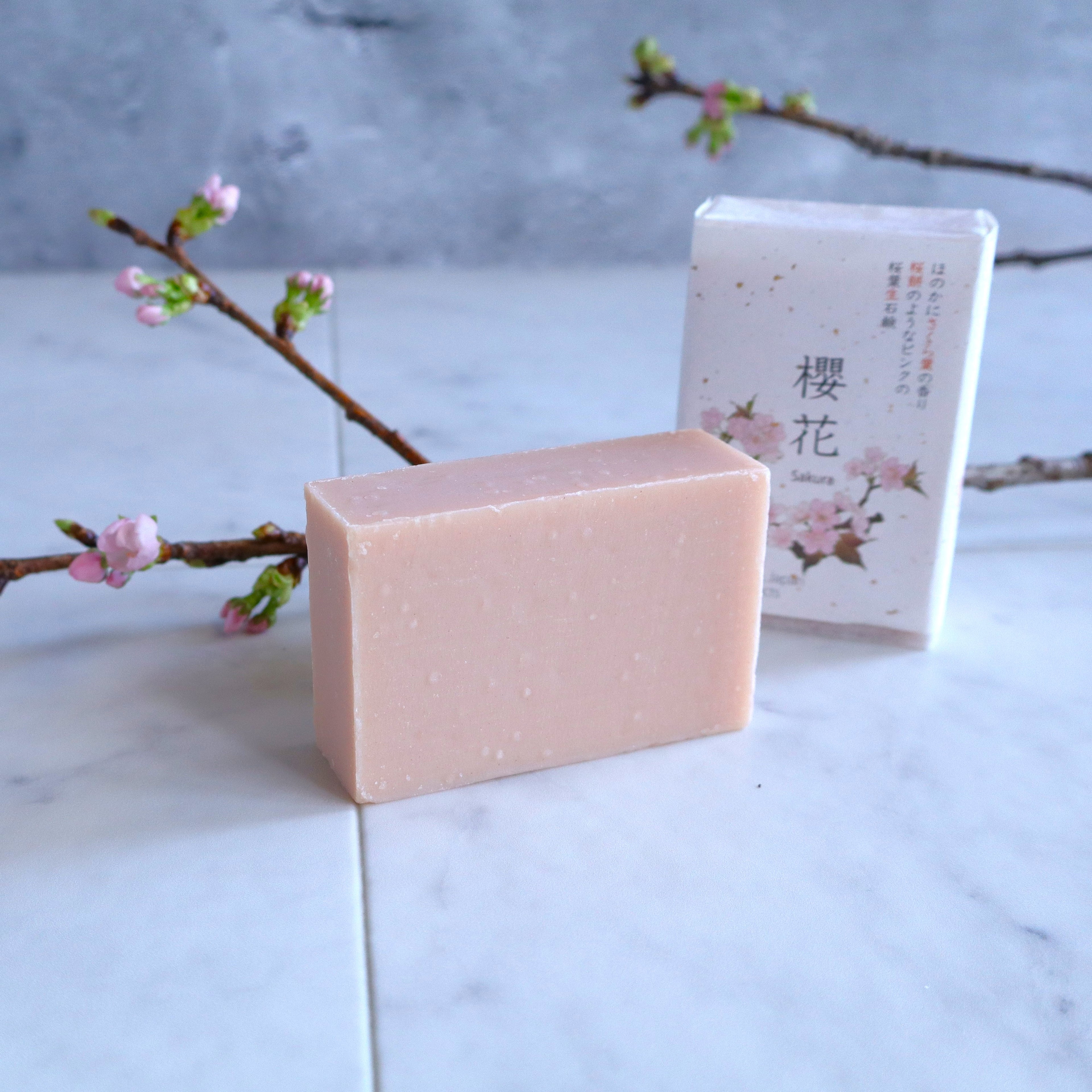 Japanese Sakura soap with a delicate pink hue, displayed alongside its elegant cherry blossom packaging on a marble surface, with blooming sakura branches in the background for a natural and serene aesthetic.