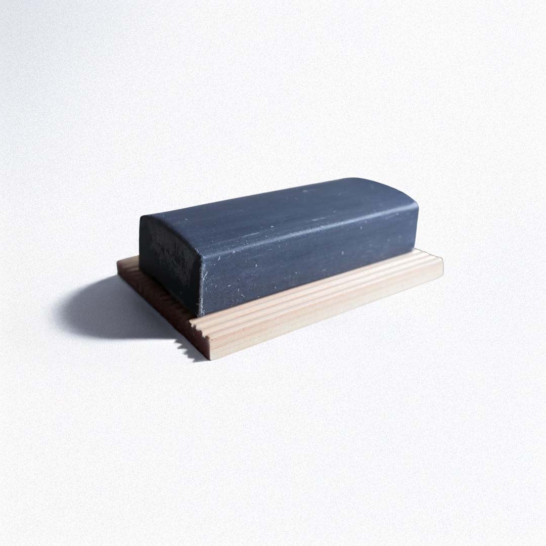 A black charcoal facial soap bar with a smooth, rectangular shape resting on a light-colored hinoki wood soap dish with a grooved design.