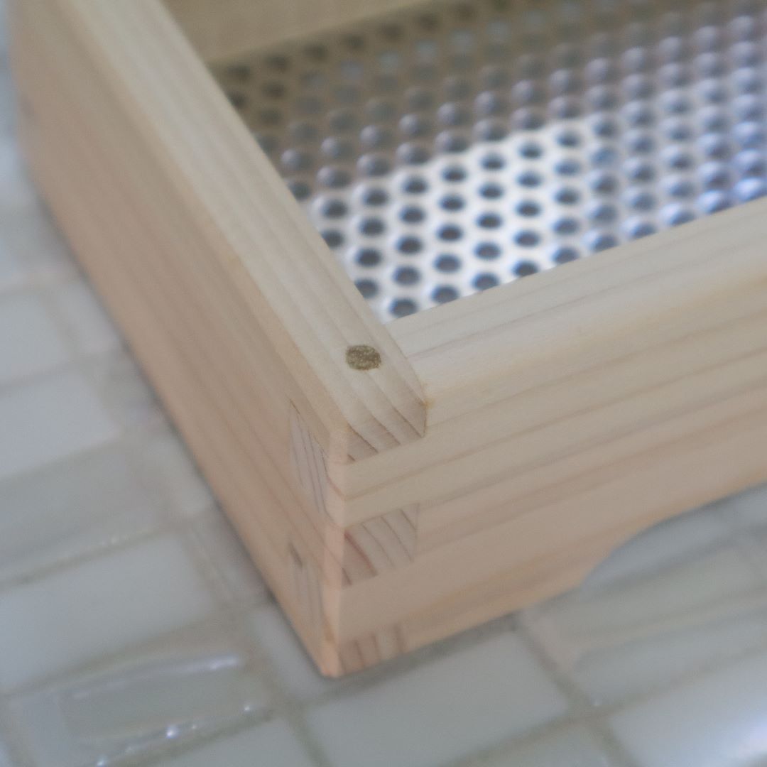A close-up of the corner of a light hinoki cypress wooden soap dish, showcasing its fine craftsmanship and joinery, set against a tiled bathroom backdrop with a perforated metal soap holder visible within.