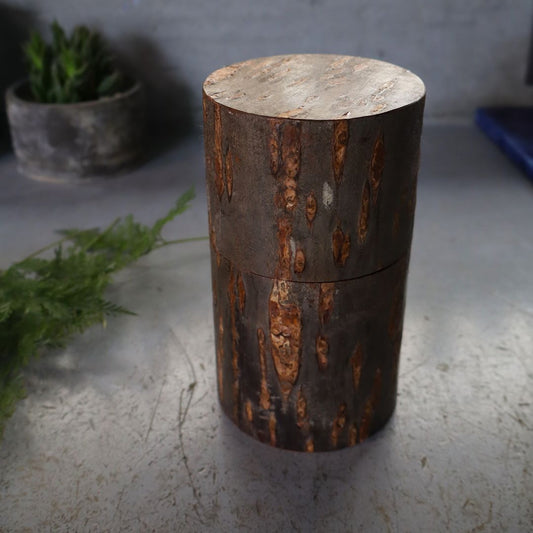 A cylindrical tea/coffee caddy made of natural wild cherry bark with a marbled exterior, displayed on a flat surface with a potted plant in the background.