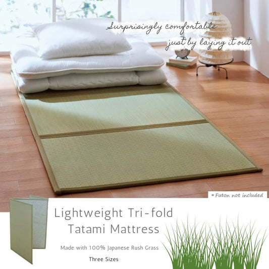 Image of a lightweight tri-fold tatami mattress laid out on a wooden floor with a fluffy white futon and pillows on top, set in a minimalist room with a large window and indoor plants, emphasizing the mattress comfort and natural material.