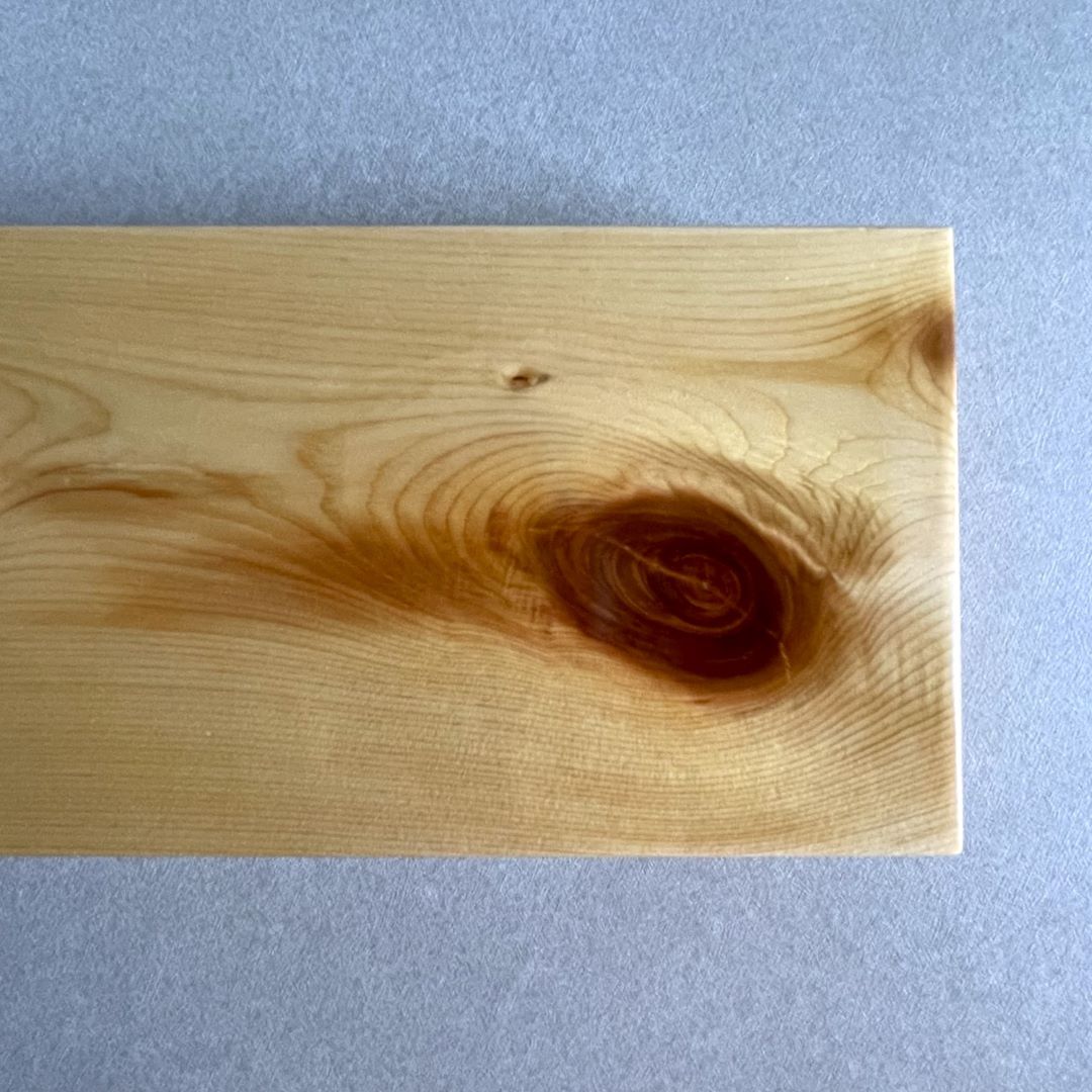 A close-up image of a section of a pine wood board showing a large, dark knot surrounded by swirling wood grain patterns. The wood&