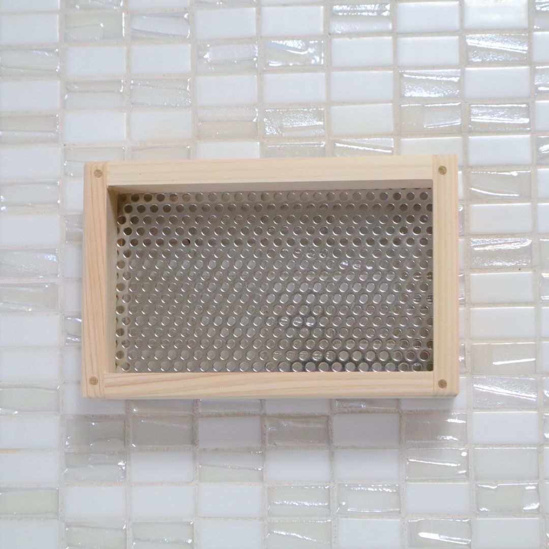 A top-down view of a rectangular hinoki cypress wooden soap dish with a perforated stainless steel panel, resting on a glossy white tiled surface, demonstrating a blend of natural wood textures and modern design.
