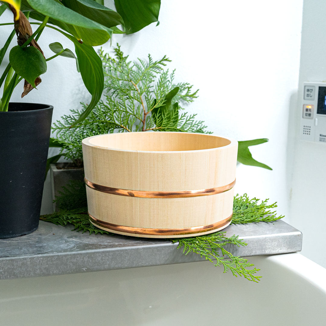A bath wooden bucket with two copper ring around it a white towel on the edge and it is placed on a granit rock with the sea on background