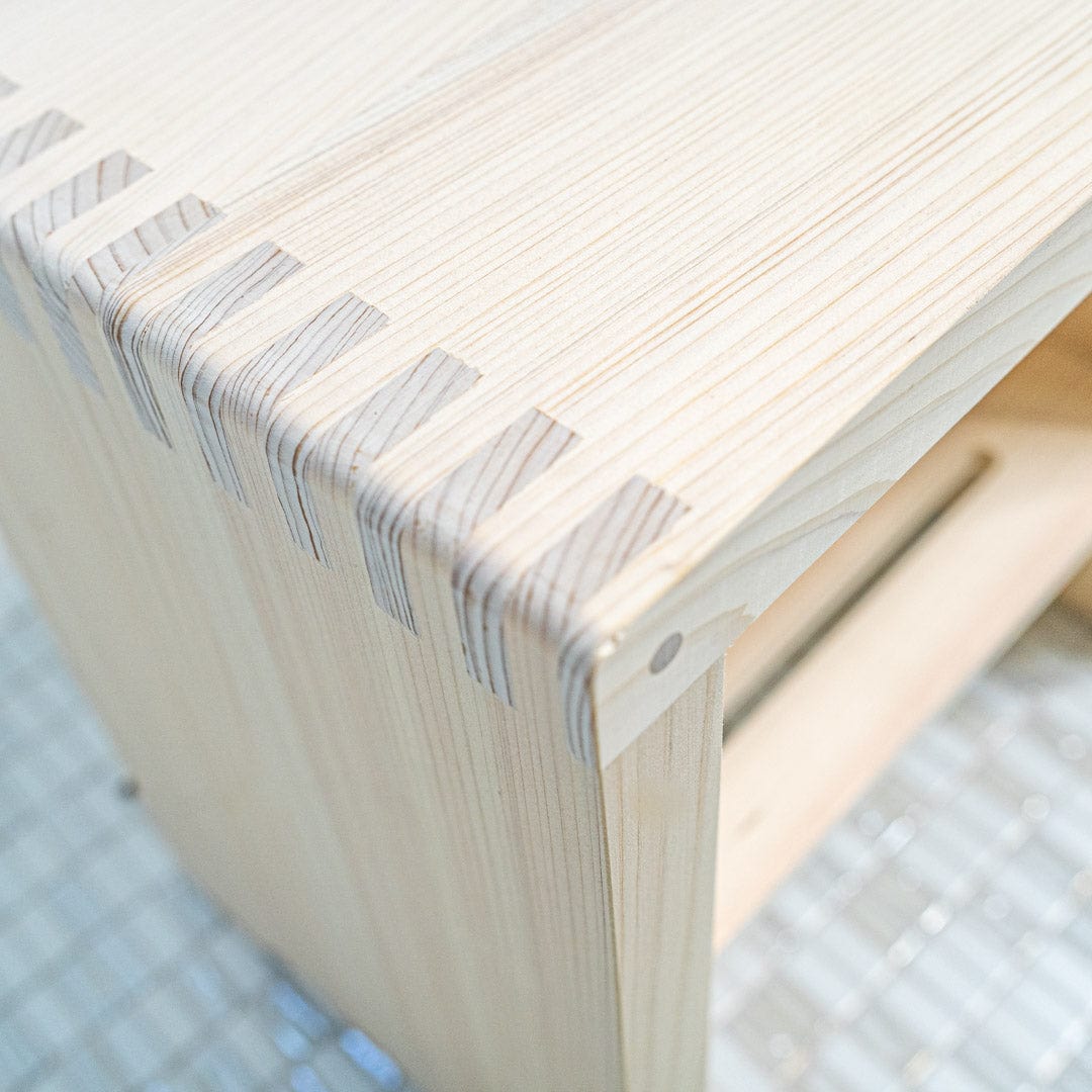 This is a close-up view of a corner of a Japanese square wooden shower stool. The natural texture of the wood is visible, with the grains and knots adding character to the stool&
