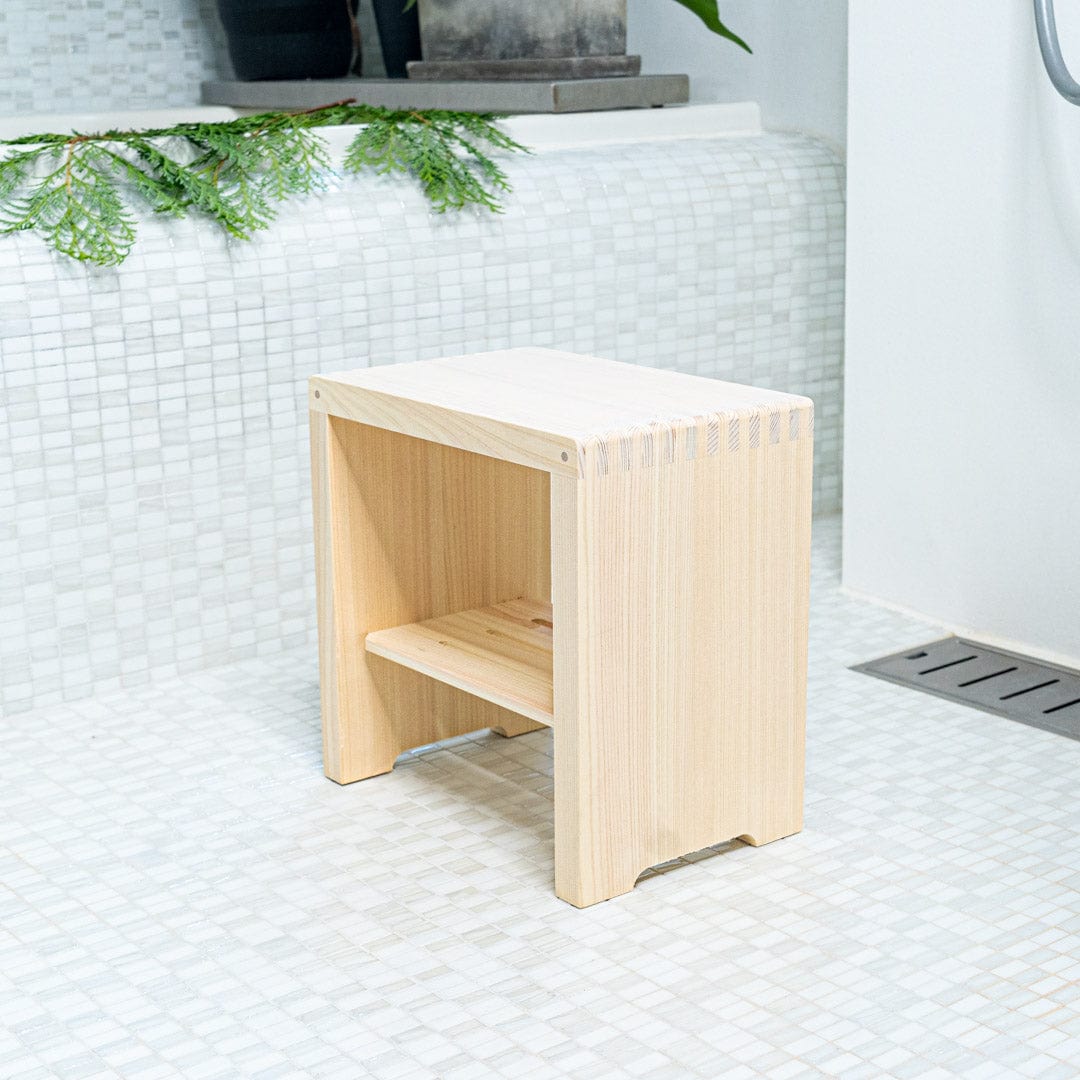 A Japanese square wooden shower stool is positioned in the center of a bathroom with faience tiling on the walls. The stool is located next to a bathtub and green plants are placed on the bathtub, adding a natural touch to the bathroom&
