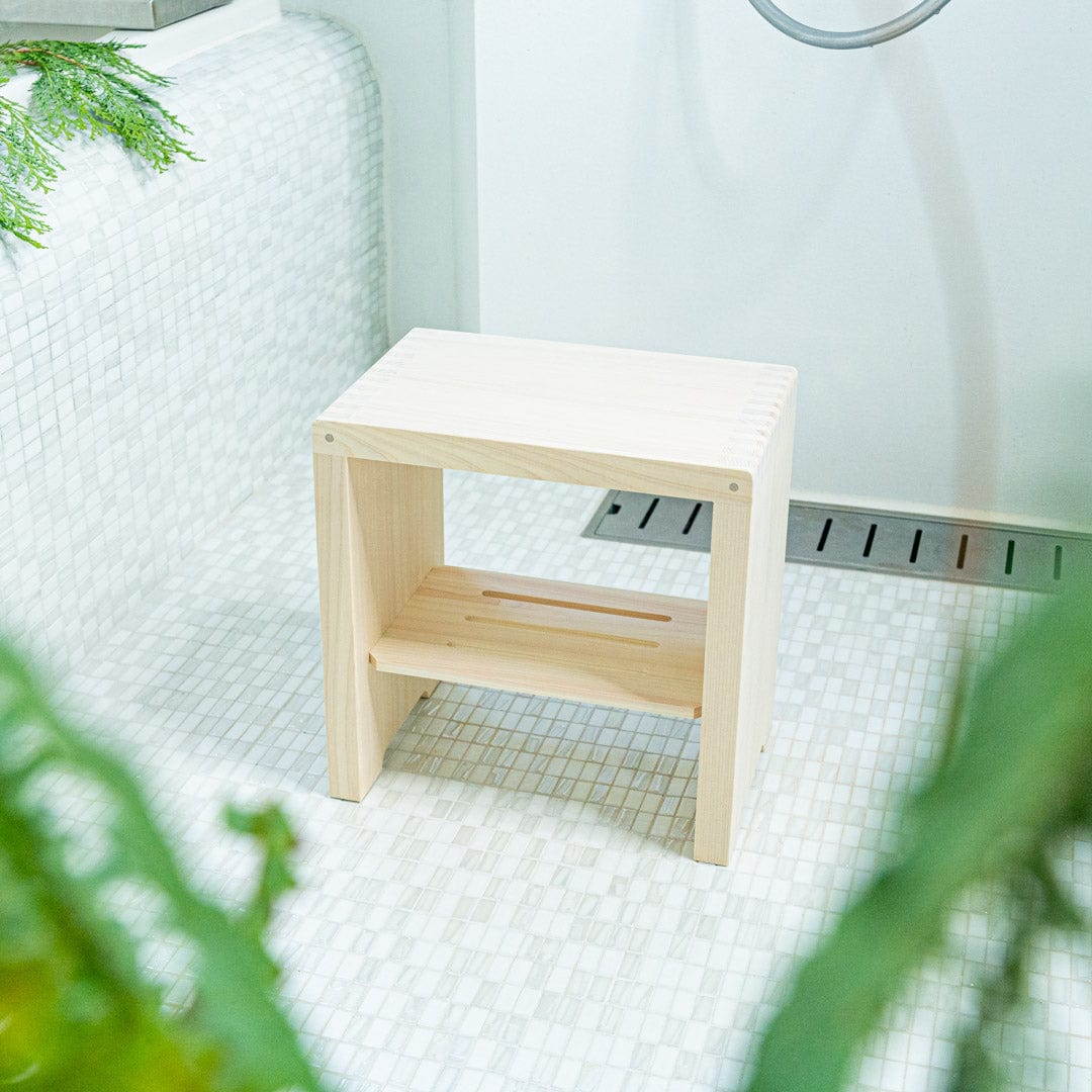 This is a view from a distance of a Japanese square wooden shower stool placed in the center of a bathroom with faience tiling on the walls. The stool is surrounded by two green plants, adding a natural element to the decor. The warm tone of the wooden stool contrasts beautifully with the cool tones of the tiles and creates a sense of harmony in the space.