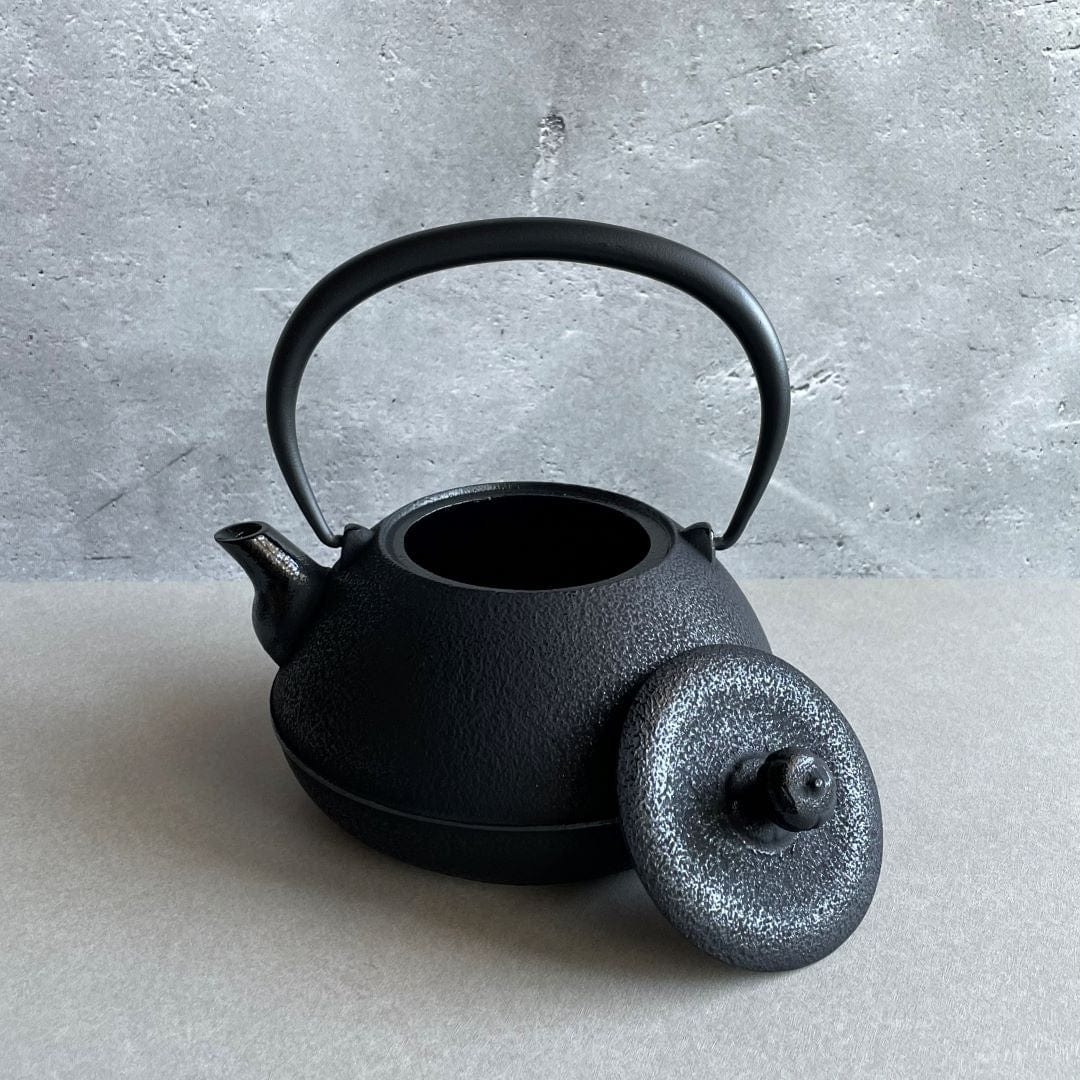 A black cast iron tetsubin kettle with lightly rough surface on the body, the iron handle has smooth surface and , the kettle is standing in the middle of a space with grey wall and grey surface, the lid is laid on the edge of the kettle.