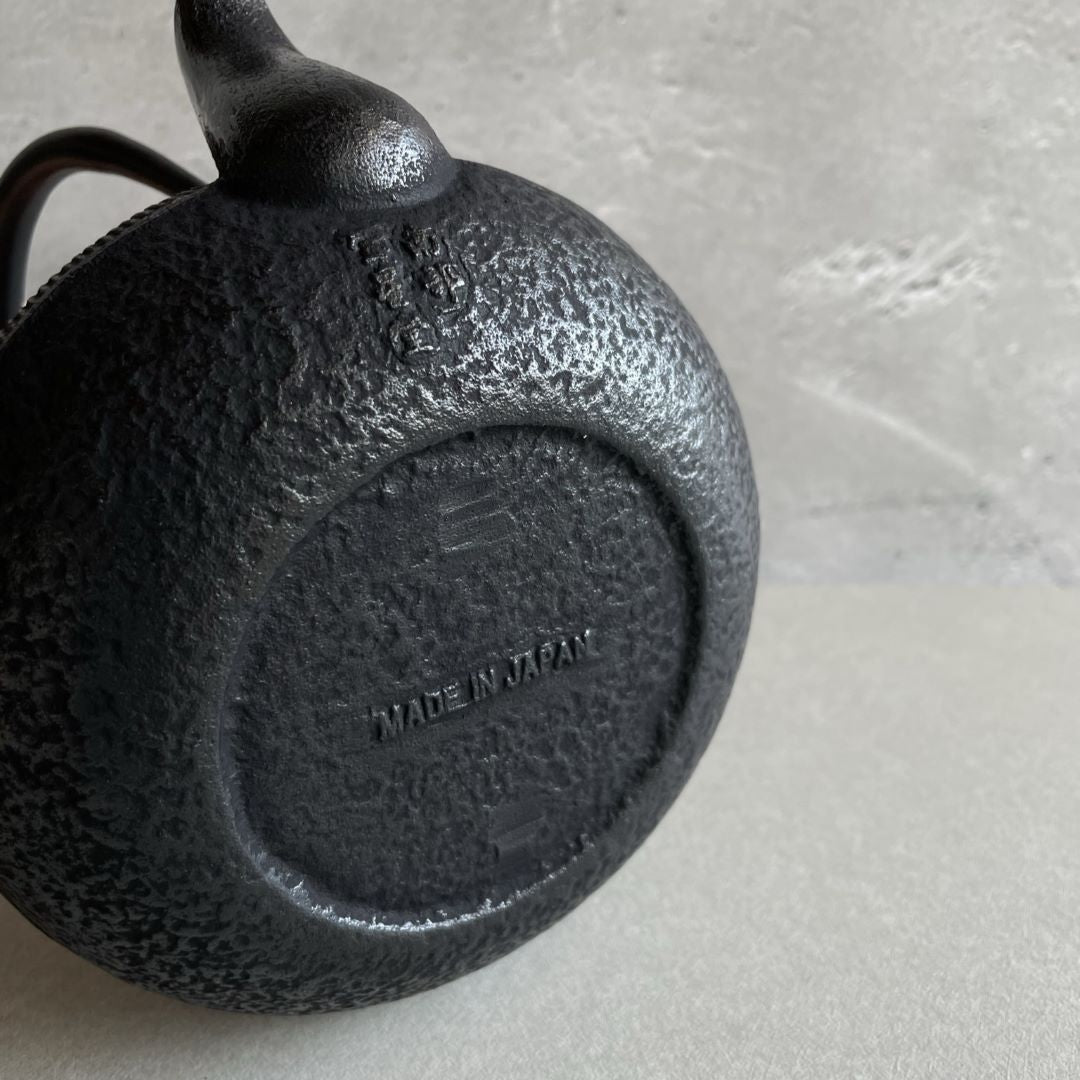 This is the underside of a black cast iron Tetsubin teapot with a rough surface. The words "Made in Japan" are written on the surface. The teapot is laid on its body, revealing the rough texture of the underside.