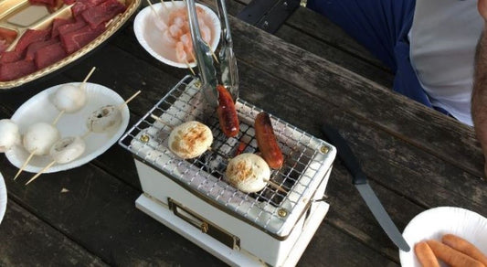 A portable grill on a wooden picnic table with sausages and mushrooms cooking on it, with plates of raw meat and shrimp nearby and a person in the background.