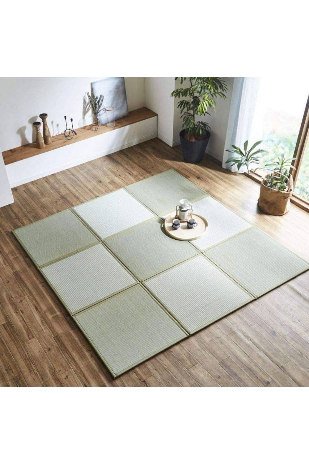 A Japanese-style room with tatami floor mats arranged in a 3x3 grid. The mats have a light green color with beige borders. In the center of the arrangement sits a small, round wooden tray with a clear glass teapot and two cups. 