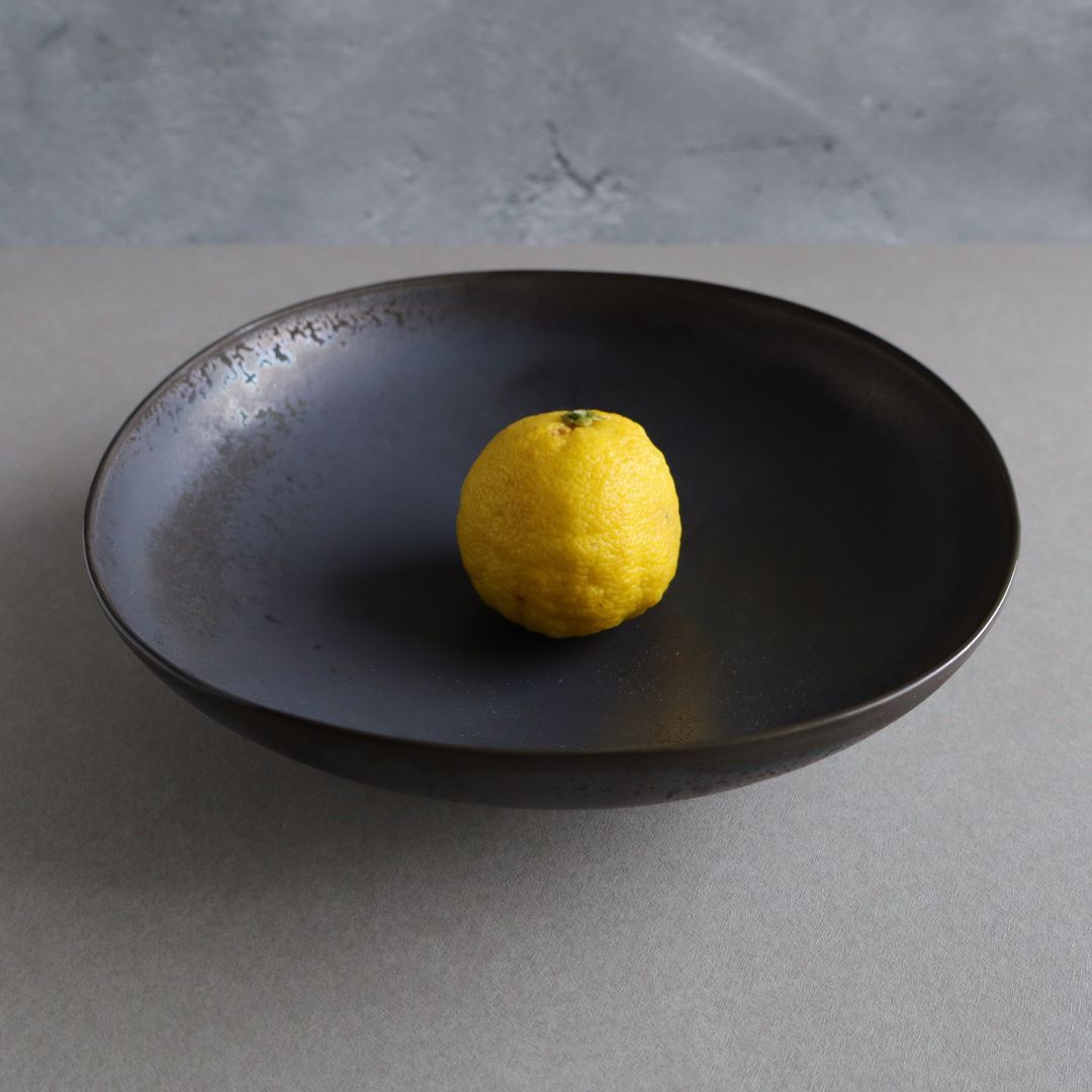 A single lemon resting on a Japanese handmade ceramic plate with a smoky charcoal finish, set against a grey background.