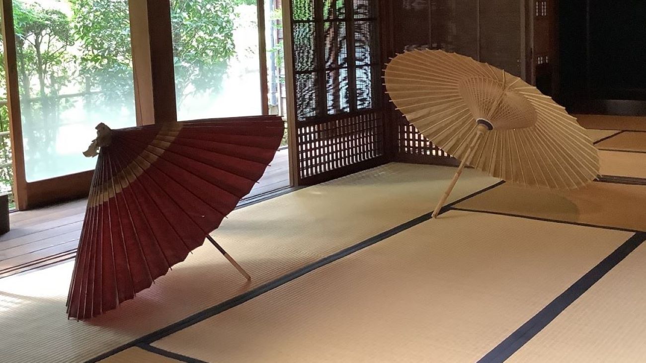 Traditional Japanese room featuring tatami flooring with two Japanese umbrellas resting on the mats. The wooden door at the back is open, revealing a view of the serene garden beyond.