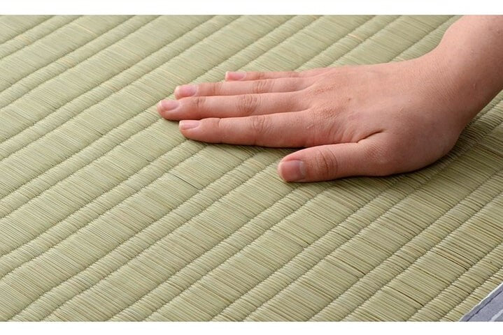 A close-up image of a hand gently placed on a tatami mat. The mat is made of woven rush grass, showcasing its texture and pattern. The hand demonstrates the smooth yet firm surface of the tatami, highlighting its quality and craftsmanship. The background is filled with the tatami mat's natural green color, providing a serene and calming visual.