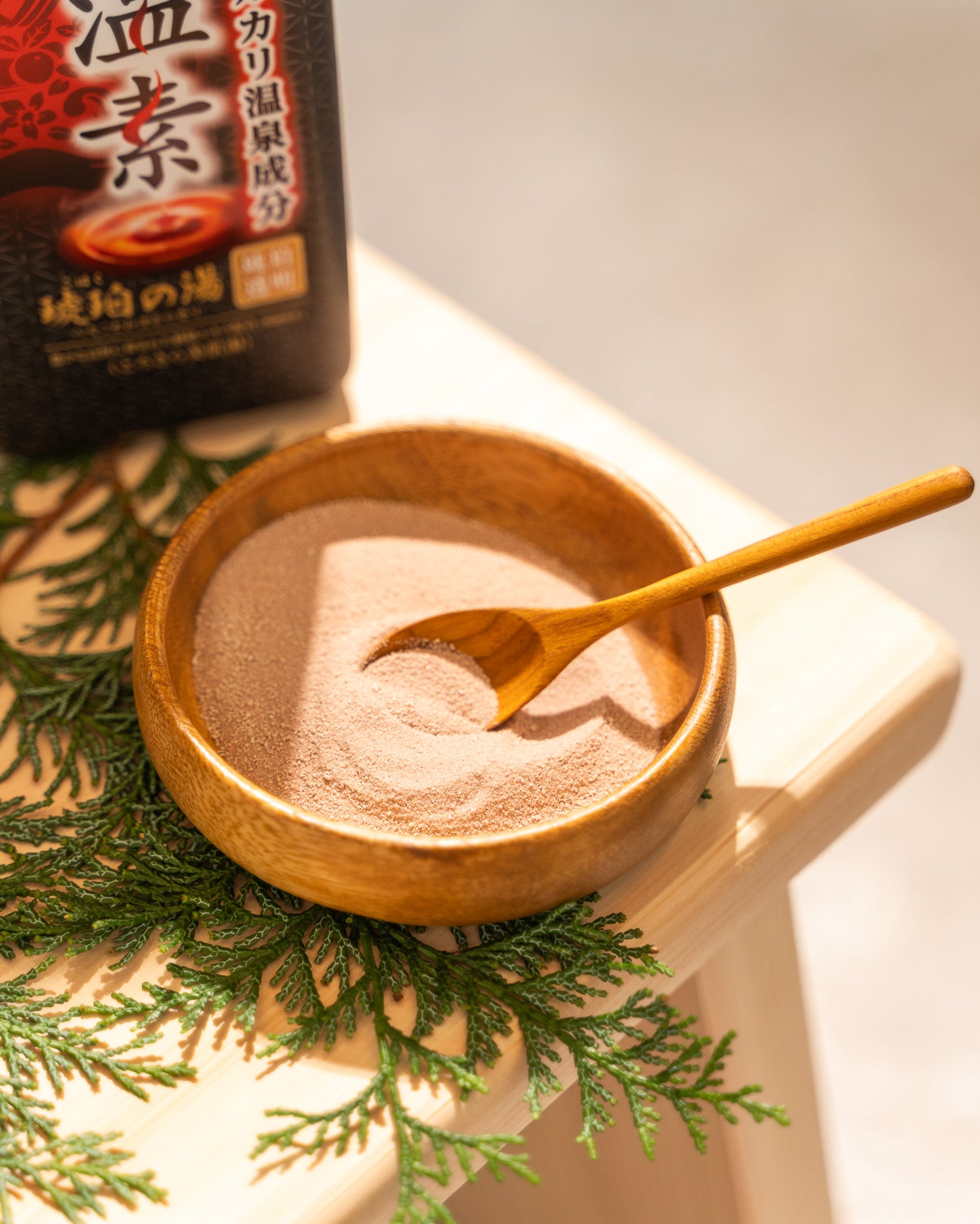 Wooden bowl with brown powder and a wooden spoon, surrounded by green leaves on a light surface.
