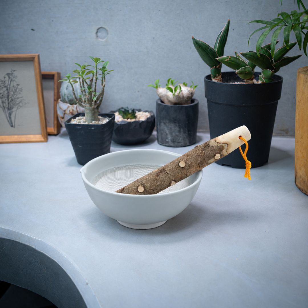 A ceramic bowl with a wooden pestle and a small orange hanging cord, styled on a light gray surface with potted plants and framed art in the background.