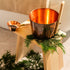 Copper sauna bucket and ladle on a hinoki wood stool decorated with fresh green cedar leaves, creating a natural spa atmosphere.
