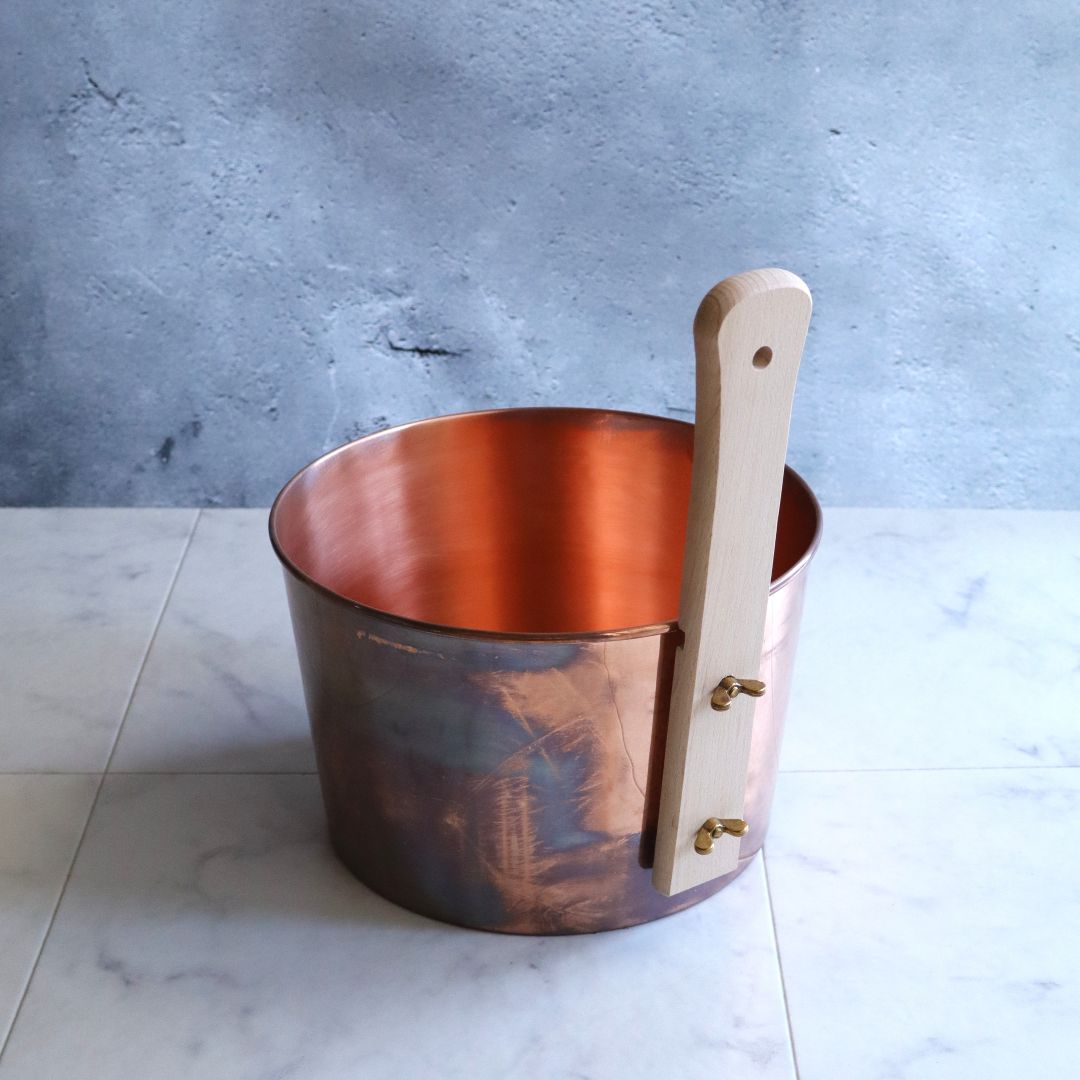 Handmade Japanese copper sauna bucket showing rear handle attachment with brass fittings, on a marble tile background.