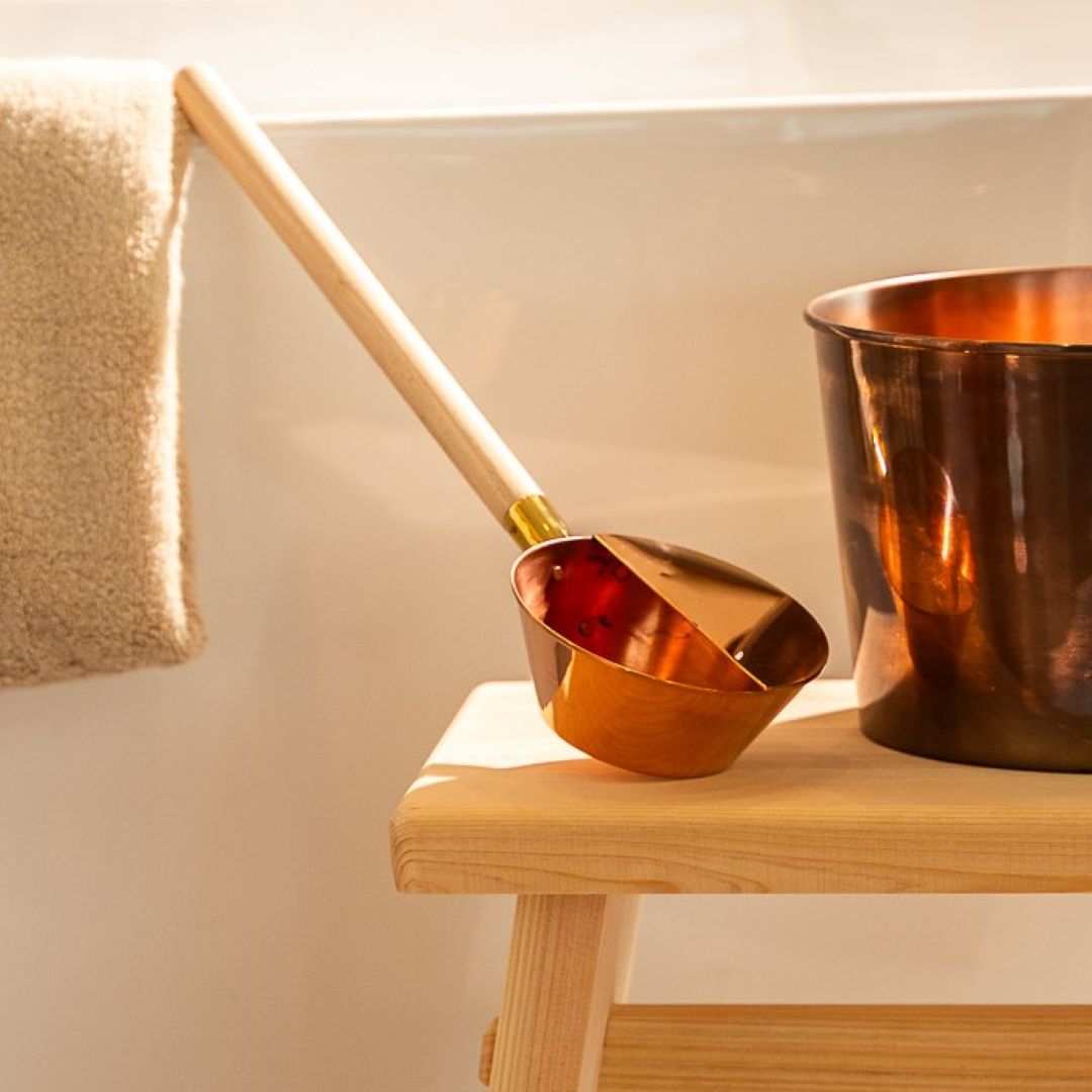 A copper sauna ladle resting on a wooden stool beside a towel, creating a warm and inviting sauna atmosphere.