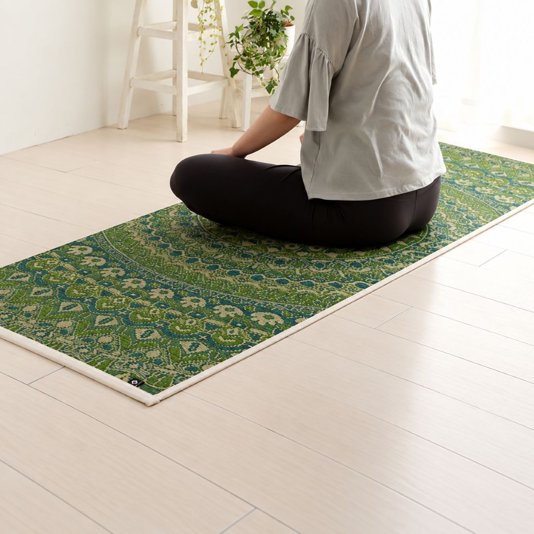 A person sits cross-legged on a green patterned yoga mat on a light wood floor in a bright room.
