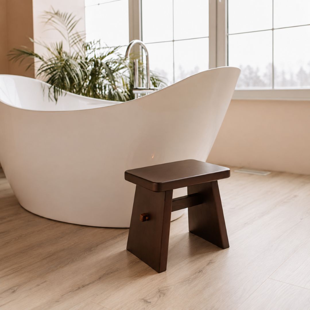 A small dark brown wooden step stool with a single knob, positioned in a bright, modern bathroom next to a white freestanding bathtub and light wood floors.