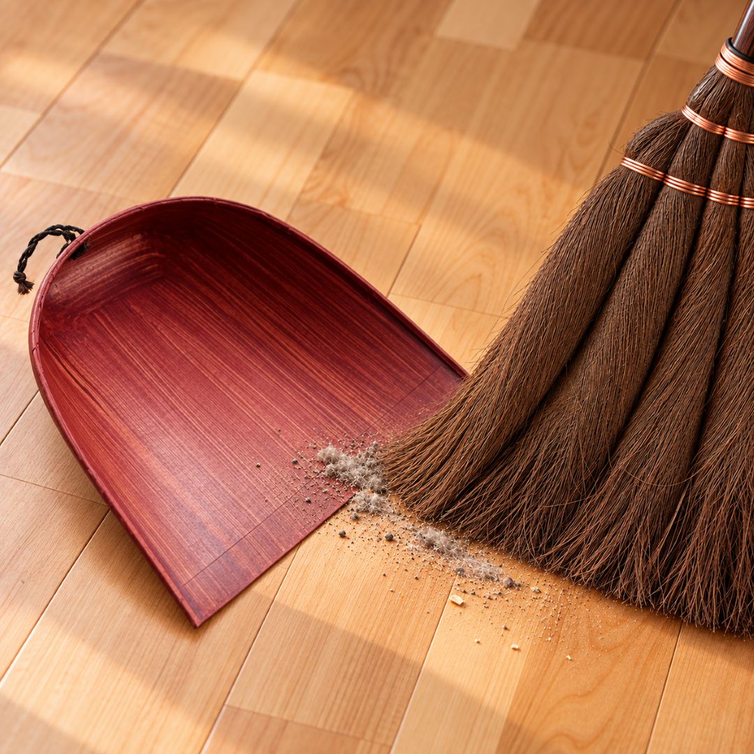 A wooden dustpan with a reddish-brown finish and a traditional brown broom with copper-colored bands, shown on a light wood floor with some dust and debris being swept.