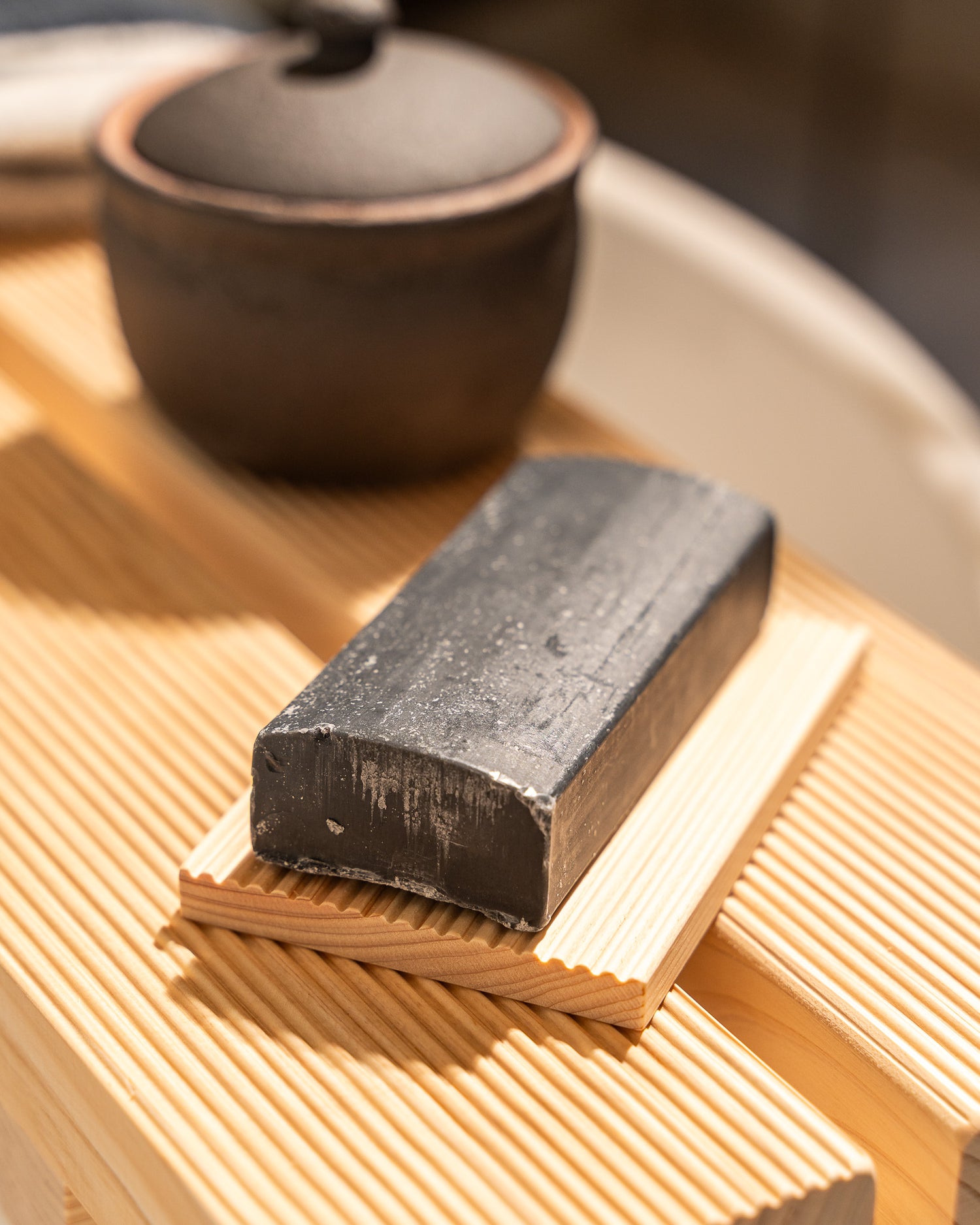 Block of dark soap on a wooden surface with a blurred teapot in the background
