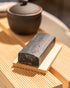 Block of dark soap on a wooden surface with a blurred teapot in the background